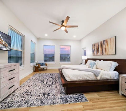 Bright contemporary master bedroom with low wood platform bed, patterned area rug, ceiling fan, dresser with TV and three windows showing a sunset view