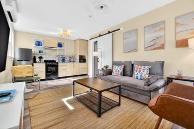Bright, cozy studio apartment open-plan living room with kitchenette, light wood cabinets, black stove and fridge, gray sofa with colorful coastal pillows, metal coffee table on woven rug and beach-print wall art