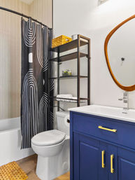 Small modern bathroom with navy blue vanity and gold hardware, white sink and toilet, black-and-white patterned shower curtain over a tub, metal over‑toilet shelving with wicker basket, and round wooden-framed mirror