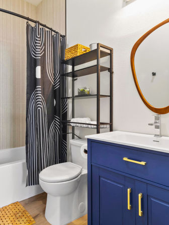 Small modern bathroom with navy blue vanity and gold hardware, white sink and toilet, black-and-white patterned shower curtain over a tub, metal over‑toilet shelving with wicker basket, and round wooden-framed mirror