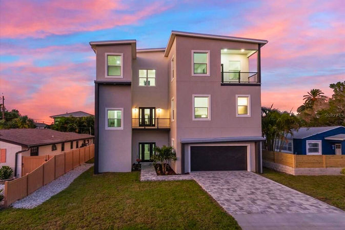 Modern three-story home with double garage and paved driveway, illuminated windows and balcony, set against a vibrant pink-and-blue sunset sky in a suburban neighborhood.