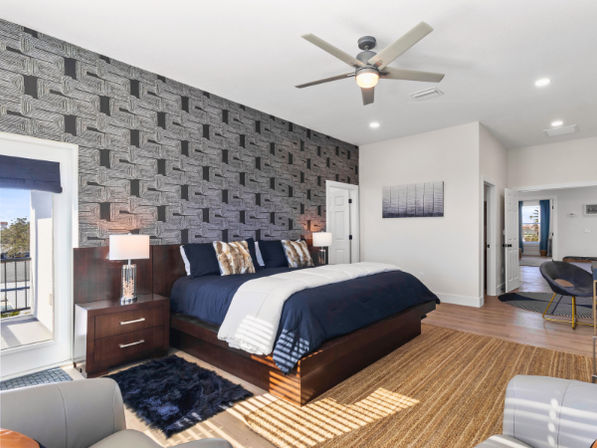 Sunlit contemporary master bedroom with navy bedding on a dark wood platform bed, geometric textured accent wall, ceiling fan, bedside lamps, balcony door, jute rug and cozy seating.