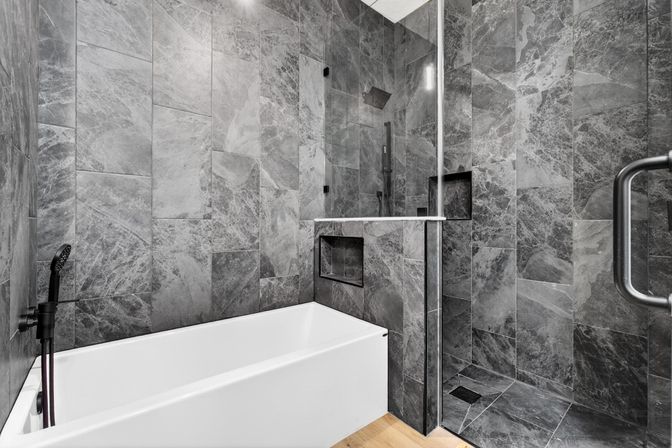 Sleek modern bathroom with large gray marble wall tiles, white soaking tub, glass-enclosed walk-in shower, matte black fixtures, recessed shower niche and warm wood floor accent.
