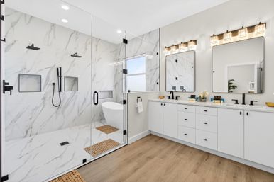Spa-like bright modern bathroom with glass-enclosed marble walk-in shower and built-in niches, freestanding soaking tub, white double vanity with black matte faucets and illuminated mirrors, and wood-look flooring.