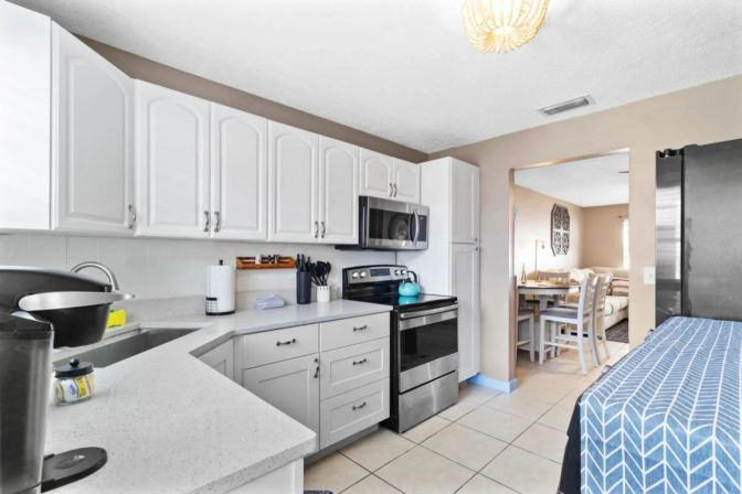 Bright modern U-shaped kitchen with white cabinets, quartz countertops, stainless steel stove and microwave, turquoise kettle on the range, tiled floor, and a dining area visible through the doorway.