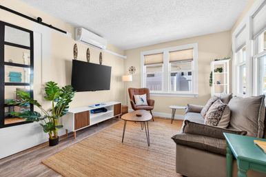Cozy modern living room with wall-mounted flat-screen TV, mid-century media console, gray sofa with patterned pillows, leather accent chair, potted tropical plant, jute area rug, and sunlit windows