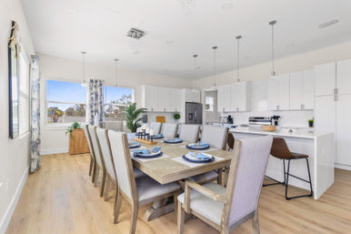 Bright open-concept modern kitchen and dining area with light wood floors, large wooden table set for eight with blue place settings, upholstered chairs, white cabinets, island with bar stools and pendant lights.