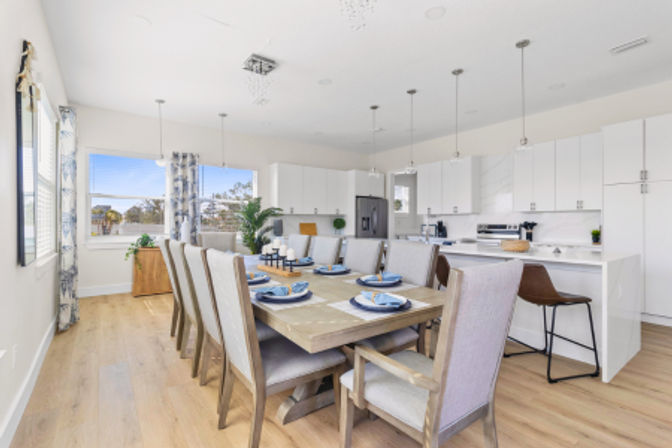 Bright open-concept modern kitchen and dining area with light wood floors, large wooden table set for eight with blue place settings, upholstered chairs, white cabinets, island with bar stools and pendant lights.