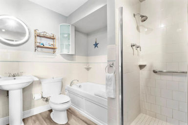 Bright coastal-style white bathroom with soaking bathtub, pedestal sink, toilet, glass-door walk-in tiled shower with grab bar, round mirror, wall shelving, towel ring, and wood-look flooring