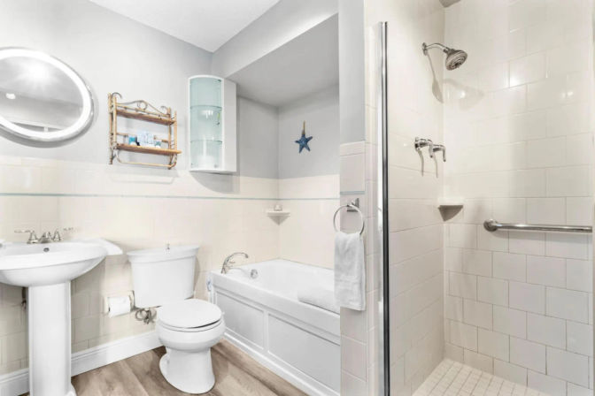 Bright coastal-style white bathroom with soaking bathtub, pedestal sink, toilet, glass-door walk-in tiled shower with grab bar, round mirror, wall shelving, towel ring, and wood-look flooring