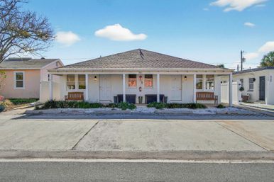 Curbside view of a single-story white duplex with two front doors and matching wooden benches, low landscaping and gravel beds beneath a wide porch on a sunny suburban street.