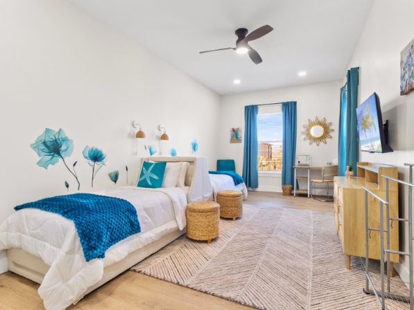 Bright coastal-style twin bedroom interior with white bedding, turquoise throws and pillows, blue floral wall decals, wicker stools, natural wood dresser and desk, teal curtains on a large window, ceiling fan and light wood flooring.