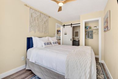 Cozy coastal-style bedroom with white bedding and pillows reading 'NO WAKE ZONE', macramé wall hanging, wooden ceiling fan, sliding barn-door closet, and wood-look floors.
