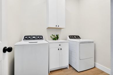 Bright modern laundry room with white top-load washer and matching dryer flanking a white storage cabinet, small countertop with a green potted plant, and light wood flooring.
