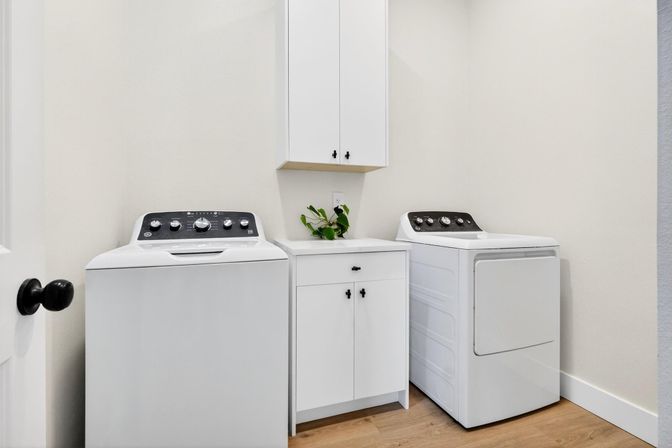 Bright modern laundry room with white top-load washer and matching dryer flanking a white storage cabinet, small countertop with a green potted plant, and light wood flooring.