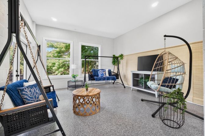 Bright sunroom lounge with hanging swing sofas and rattan egg chair, blue cushions, round carved wood coffee table, TV console and large windows overlooking green trees
