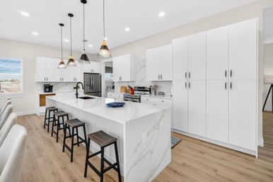 Modern open-concept kitchen with white cabinets, marble waterfall island, black pendant lights, wood bar stools, and stainless steel appliances on light hardwood floors.