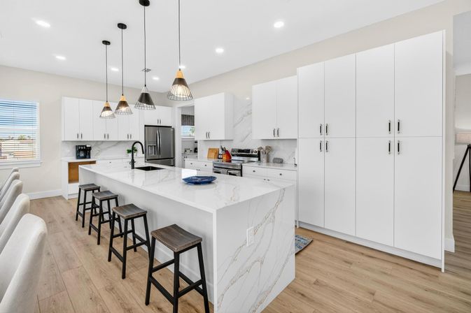 Modern open-concept kitchen with white cabinets, marble waterfall island, black pendant lights, wood bar stools, and stainless steel appliances on light hardwood floors.