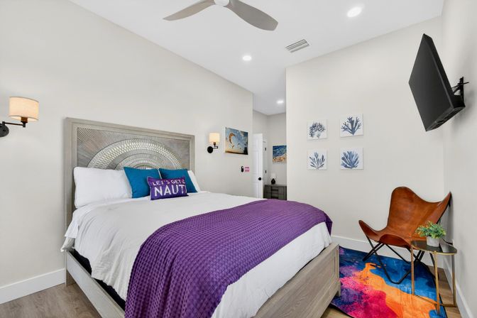 Bright coastal-themed vacation rental bedroom with queen bed, decorative sunburst headboard, white linens, purple throw and “Let's Get Naut” accent pillow, wall-mounted TV, leather butterfly chair, coral artwork and colorful area rug