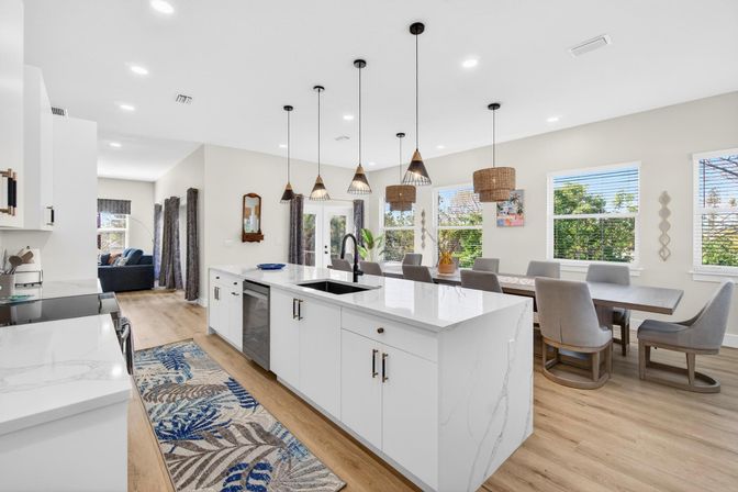 Sunlit modern open-concept kitchen and dining area with a white marble island, black faucet, mixed pendant lights, wood floors, large windows and upholstered dining chairs.