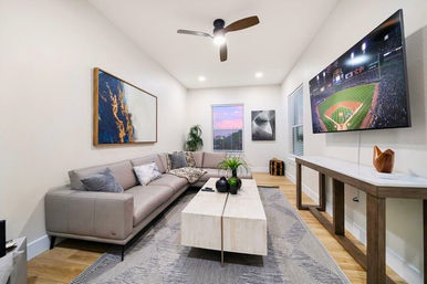 Bright modern living room with a sleek gray leather sectional, light wood coffee table, wall-mounted TV showing a baseball game, abstract wall art, hardwood floors, area rug, ceiling fan, and a window with a pink sunset.