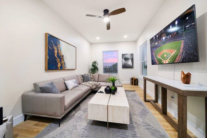 Bright modern living room with a sleek gray leather sectional, light wood coffee table, wall-mounted TV showing a baseball game, abstract wall art, hardwood floors, area rug, ceiling fan, and a window with a pink sunset.