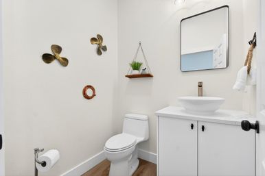 Bright modern residential powder room with white toilet and cabinet, round vessel sink, black-framed mirror, floating wood shelf with potted plant, nautical propeller wall decor and wood-look flooring