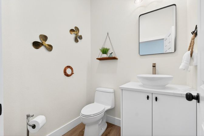 Bright modern residential powder room with white toilet and cabinet, round vessel sink, black-framed mirror, floating wood shelf with potted plant, nautical propeller wall decor and wood-look flooring