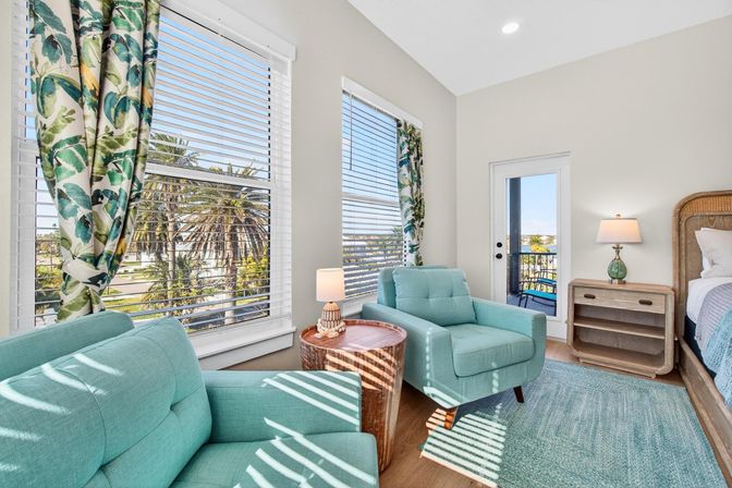 Sunlit coastal bedroom with teal armchairs, tropical-print curtains and palm trees visible through wide blinds, wooden side tables, rattan headboard, and a balcony door with ocean view.