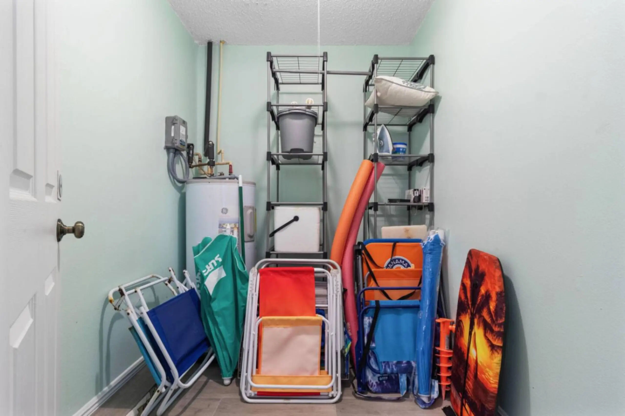 Small home utility closet with a water heater, metal shelving and stacked beach gear—folding beach chairs, colorful pool noodles, a palm-sunset boogie board, bucket and pillow.