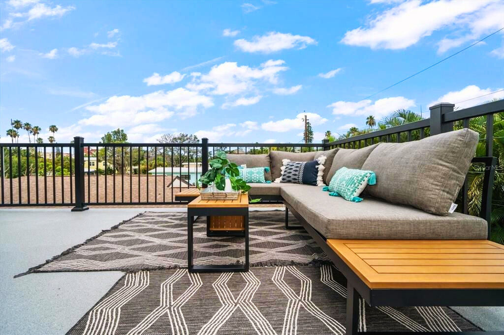Sunny rooftop patio lounge with gray sectional sofa, teal accent pillows, wooden side tables and coffee table, patterned outdoor rugs, potted plant, black metal railing and palm trees under a bright blue sky.