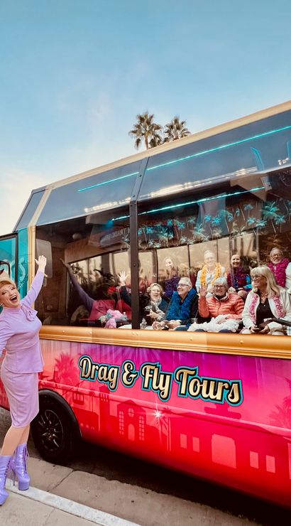Drag performer in lavender outfit and platform boots poses beside a bright pink tour shuttle; inside, a festive group wearing leis and boas waves, with palm trees and a sunset sky reflected on the windows.