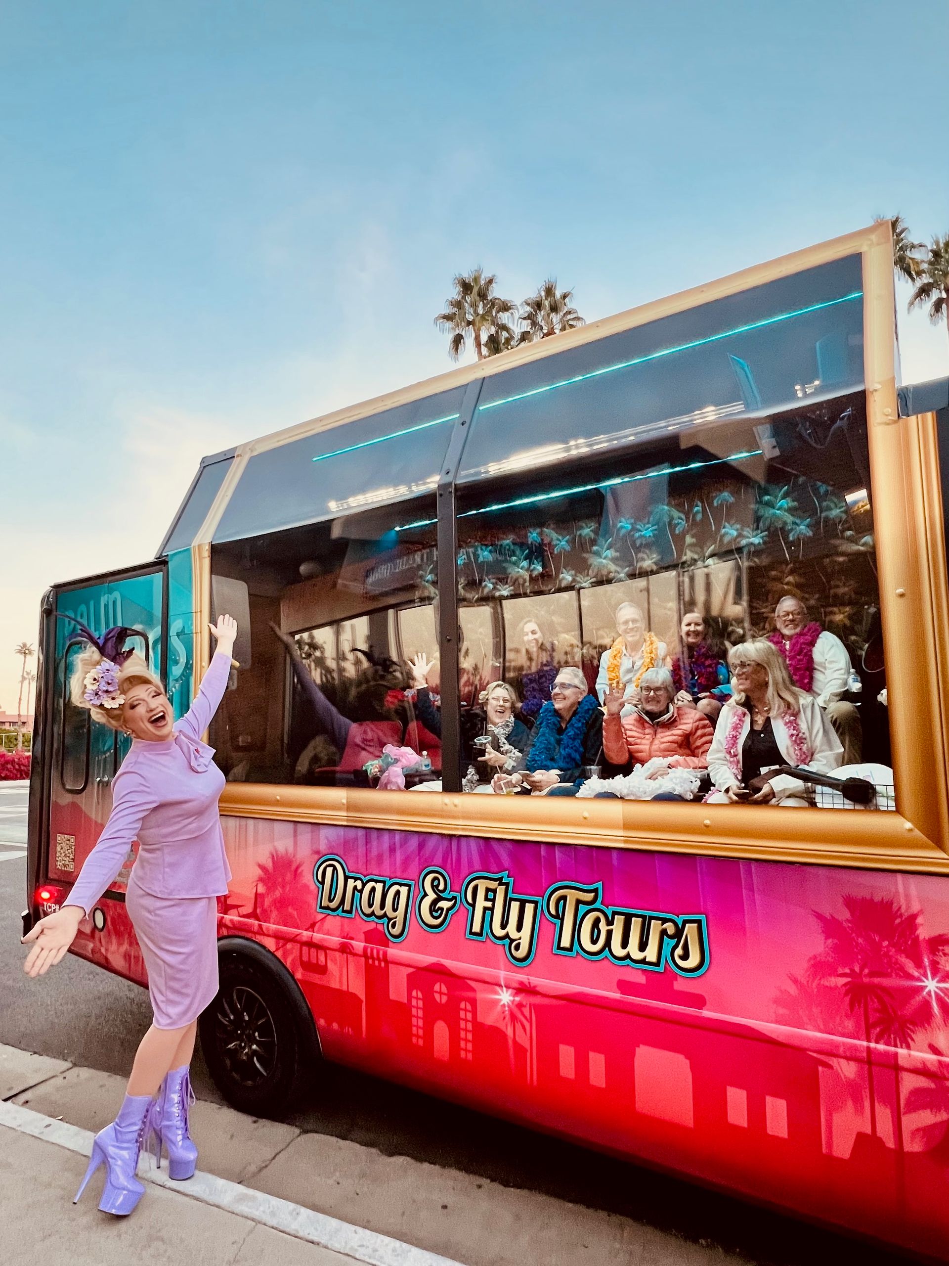Drag performer in lavender outfit and platform boots poses beside a bright pink tour shuttle; inside, a festive group wearing leis and boas waves, with palm trees and a sunset sky reflected on the windows.