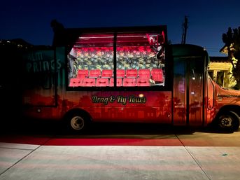 Red tour bus parked at night in Palm Springs, neon-pink lit interior showing rows of red velvet seats and tropical palm-leaf wallpaper, silhouetted palms against a deep blue evening sky.