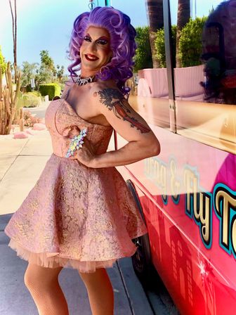 Drag performer in a lavender wig and glittery pink brocade dress posing by a pink food truck on a sunny desert street lined with palm trees and cacti.