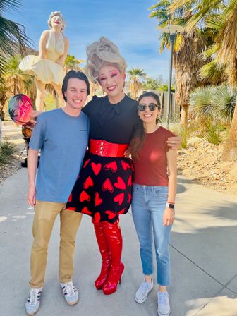 Smiling couple posing with a tall drag performer in a black skirt patterned with red hearts, wide red belt and shiny red knee-high boots on a sunny, palm-lined walkway with a large blonde statue in the background.