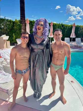 Poolside photo of a purple‑wigged drag performer in a shimmering silver gown posing between two shirtless men in colorful swim briefs by a palm tree and turquoise pool on a sunny day.
