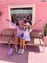 Two smiling people posing on a pink bench outside a pastel pink storefront with neon script — one in a lavender outfit, pink wig and fringed heels, the other in a white dress and wide-brimmed hat.