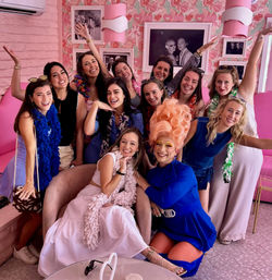 Group of smiling women and a drag performer in a towering pink wig posing together in a bright pink, floral-themed cafe for a fun girls’ brunch photo.