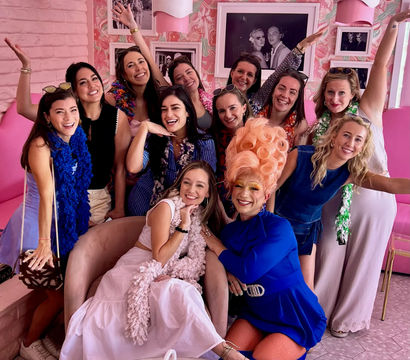 Group of smiling women and a drag performer in a towering pink wig posing together in a bright pink, floral-themed cafe for a fun girls’ brunch photo.