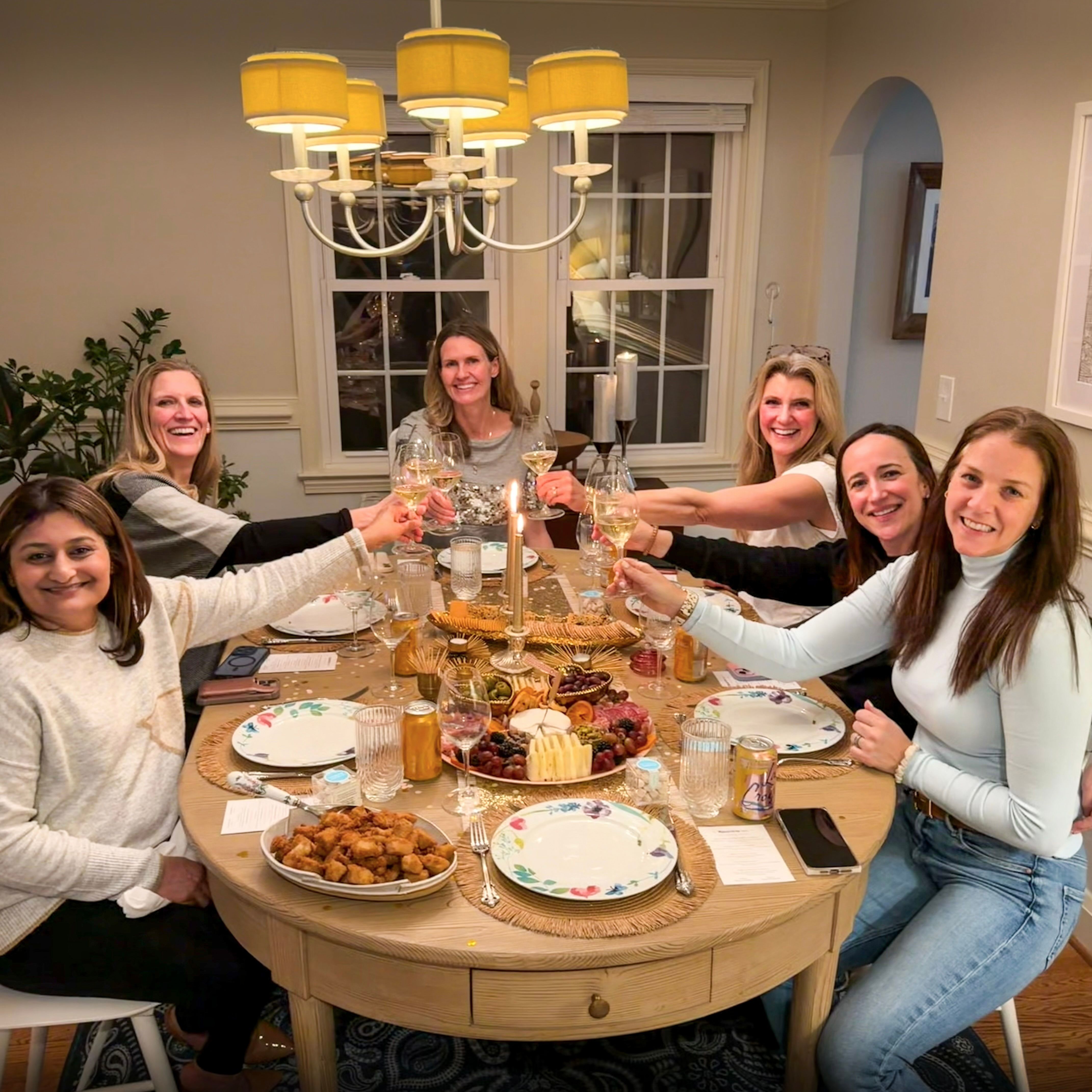 Six women at a cozy home dinner party toasting with white wine around a round table set with candles, plates, and a cheese and charcuterie board.