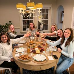 Six women at a cozy home dinner party toasting with white wine around a round table set with candles, plates, and a cheese and charcuterie board.