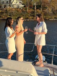 Three women in light dresses laughing and holding wine glasses on a yacht at a lakeside sunset, with a lakefront house and autumn trees in the background.
