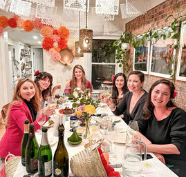Six friends smiling and toasting with wine at a cozy indoor dinner party around a decorated table with flowers, wine bottles, hanging papel picado banners, tissue pom-poms, a brick accent wall and trailing plants.