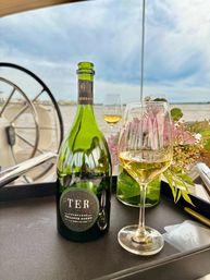 Green champagne bottle and filled wine glass on a black tray with pink floral arrangement, set on a boat deck near the helm overlooking a river under a cloudy sky.