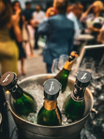 Three champagne bottles chilling in an ice bucket at a lively outdoor reception, blurred guests mingling in the background — celebration drinks station.