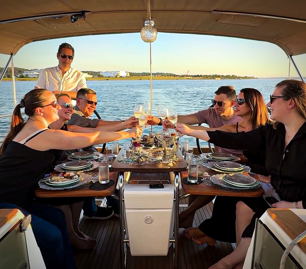 Friends toasting with wine around a dinner table on a yacht during a sunset cruise, coastal harbor and shoreline in the background.