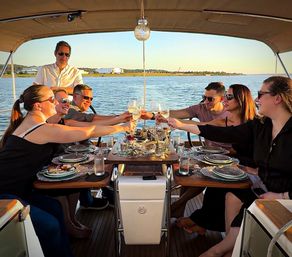 Friends toasting with wine around a dinner table on a yacht during a sunset cruise, coastal harbor and shoreline in the background.