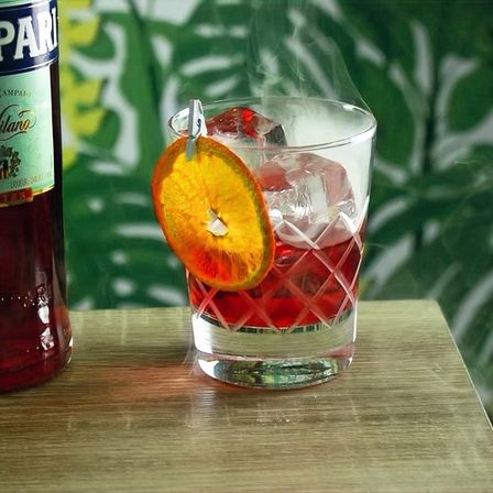 Red aperitif cocktail in a cut-crystal old-fashioned glass with large ice cubes, a dried orange wheel clipped to the rim and wisps of smoke rising, set beside a red liqueur bottle on a wooden table with a green palm-leaf backdrop.