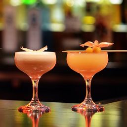 Two orange craft cocktails in coupe glasses on a bar counter — one with foam and a citrus peel, the other garnished with physalis berries, warm bokeh lights behind.